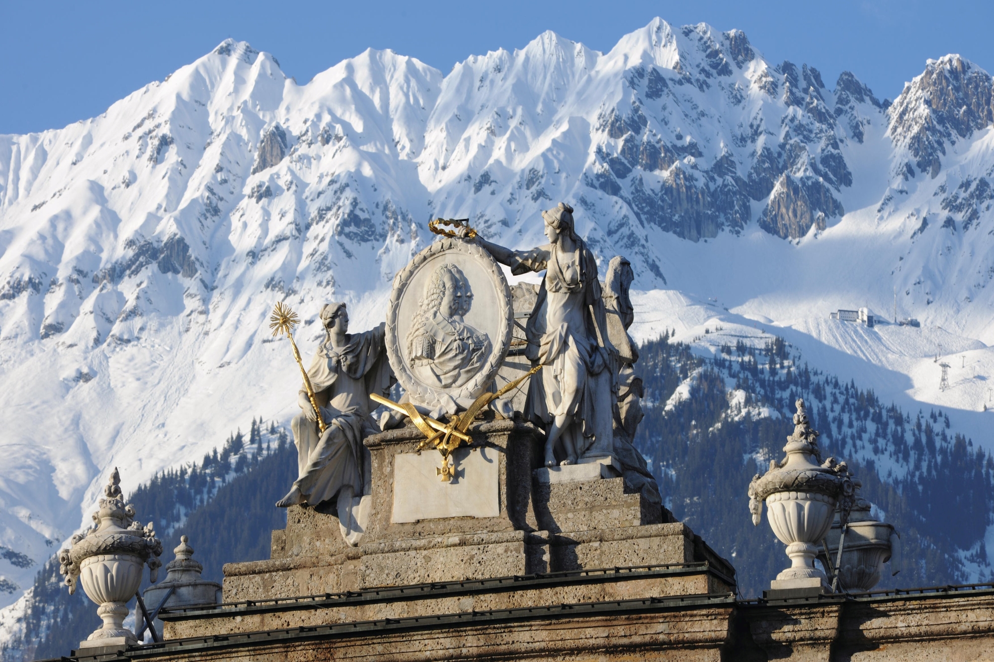 Historische Skulpturen vor schneebedeckten Bergen unter strahlend blauem Himmel.